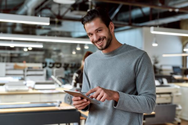 Portrait of handsome cheerful male worker in casual wear using his tablet pc and looking at camera with a smile while standing in co-working space. Horizontal shot. Selective focus