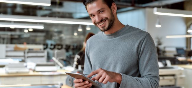 Portrait of handsome cheerful male worker in casual wear using his tablet pc and looking at camera with a smile while standing in co-working space. Horizontal shot. Selective focus