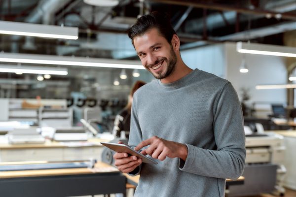 It is never too late to start afresh. Young businessman posing in office Portrait of handsome cheerful male worker in casual wear using his tablet pc and looking at camera with a smile while standing in co-working space. Horizontal shot. Selective focus