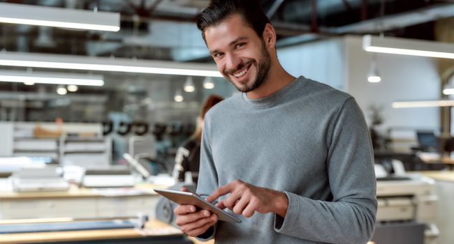 Portrait of handsome cheerful male worker in casual wear using his tablet pc and looking at camera with a smile while standing in co-working space. Horizontal shot. Selective focus