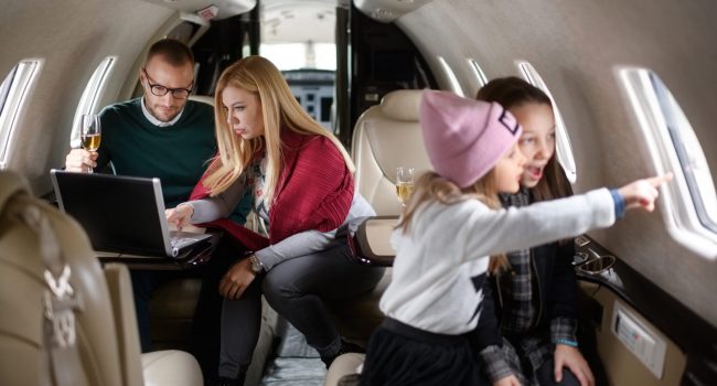 Family with two daughters sitting inside private jet airplane. Girls are playing in the front view while parents are sitting in the gackground.