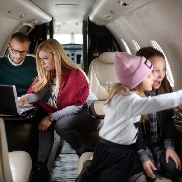 Family with two daughters sitting inside private jet airplane. Girls are playing in the front view while parents are sitting in the gackground.